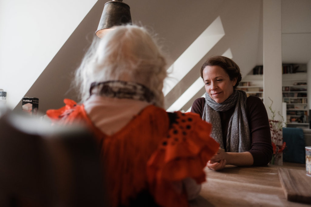 eine Mutter sitzt mit ihrer Tochter an einem großen Küchentisch, sie sitzen sich gegenüber, sie spielen ein Spiel, hinter der Mutter sieht man eine Dachschräge, in der Fenster einegesetzt sind, durch die das Licht in die Wohnung strahlt