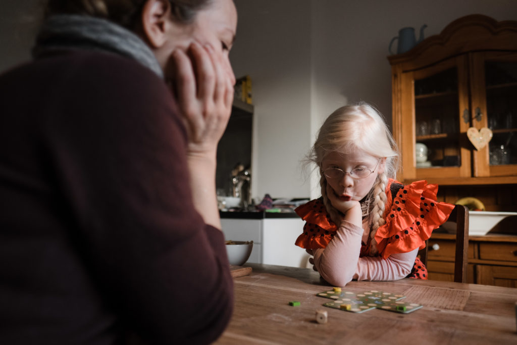 eine Muter sitzt mi ihrer Tohter an einem Küchentisch, sie spielen ein Spiel und sitzen sich dabei gegenüber, die Tochter schaut konzentriert auf das Spiel, sie stütt sich dabei ihren Kopf ab, die Mutter schaut auch konzentriert auf das Spiel