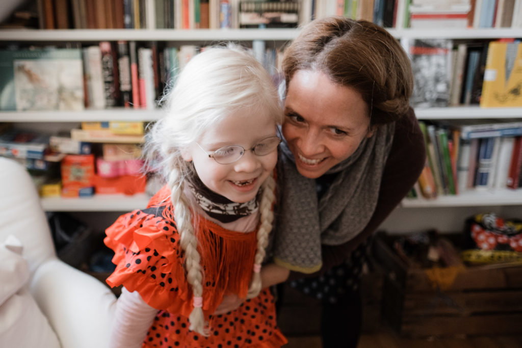 eine Mutter steht mit ihrer Tochter in einem Wohnzimmer vor einem großen Bücherregel, die Tochter trägt ein rotes Kleid, 2 Zöpfe und eine Brille, ihre Mutter steht hinter ihr und beugt sich über die Schulter der Tochter an den Kopf der Tochter