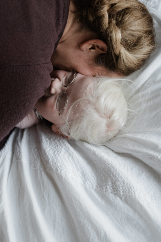 eine Mutter hält ihre Tochter in dem Armen, sie liegen dabei auf einer weißen Decke, die Mutter hält ihren Kopf an den Kopf der Tochter