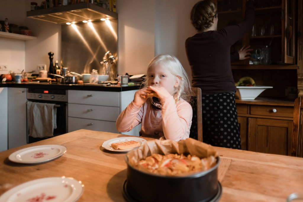 ein Mädchen sitzt an einem großen Küchentisch, auf dem Küchentich stehen 3 gemusterte Kuchenteller und ein gebackener Apfelkuchen in einer Springform, das Mädchen hält eine Waffel an den Mund, hinter ihr steht ihre Muter, die etwas aus dem Holzschrank holt