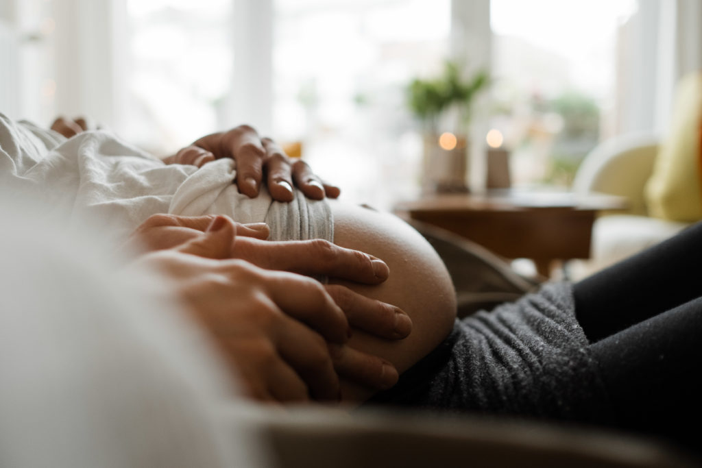 ein Mann und eine Frau liegen auf einem Sofa in einem Wohnzimmer, sie liegt in den Armen des Mannes, sie ist schwanger, die Hände des Mannes und der Frau liegen auf ihrem Bauch, nur der Bauch ist zu sehen