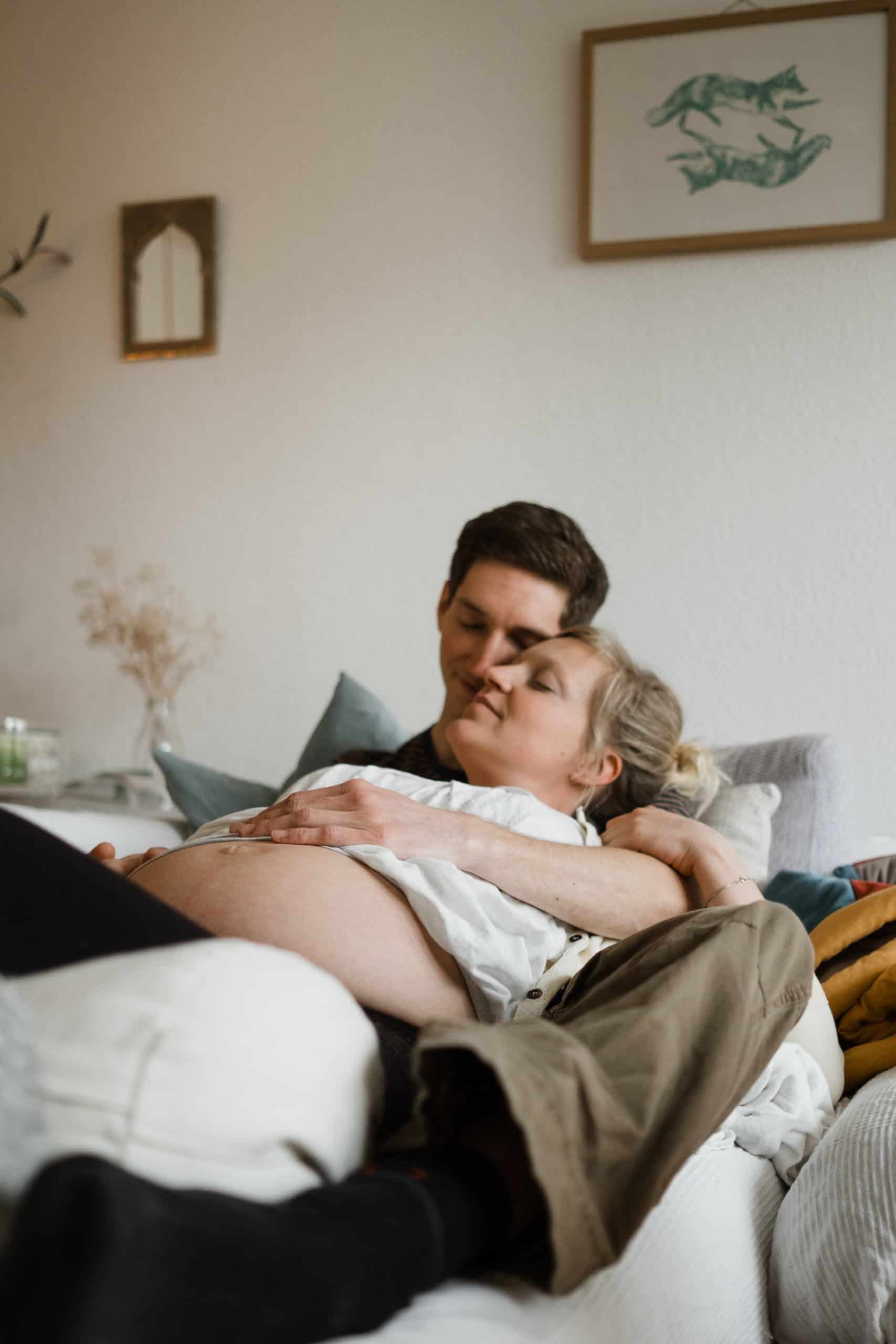 ein Mann und eine Frau liegen auf einem Sofa in einem Wohnzimmer, die Frau ist schwanger, sie liegt in den Armen des Mannes, beide haben geschlossene Augen