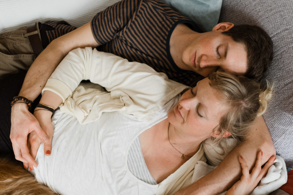 eine Mann und eine schwangere Frau liegen auf einem Sofa in einem Wohnzimmer, der Mann legt seine Arme um die Frau, beide haben geschlossene Augen