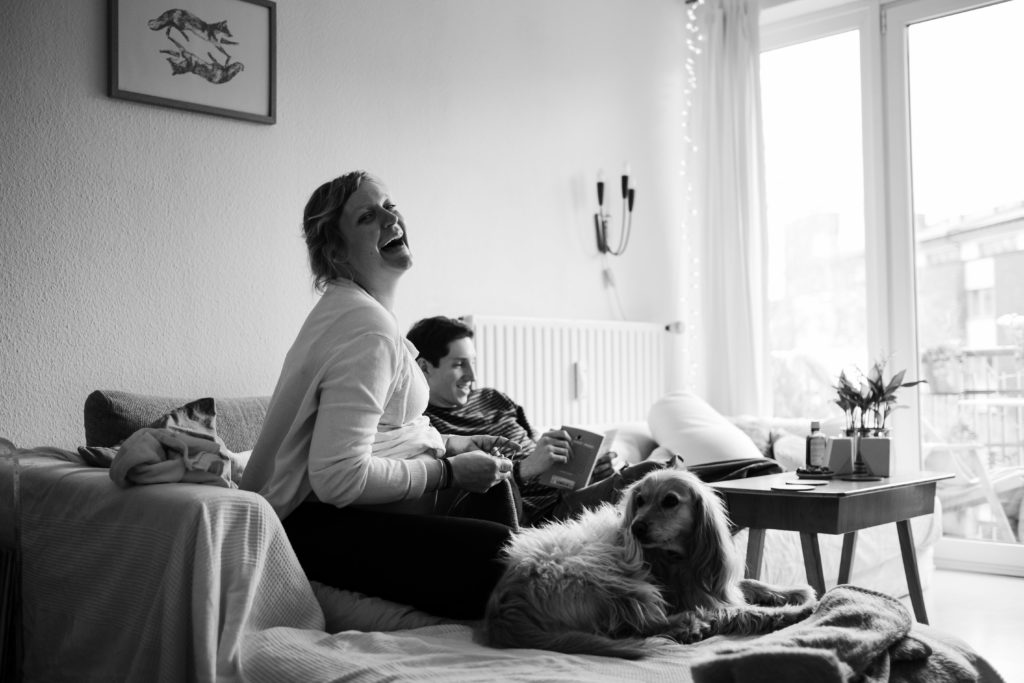 ein Mann und eine schwangere Frau sitzen auf einem Sofa in einem Wohnzimmer, die Frau ist schwanger, sie strickt, sie schaut lachend zu Seite, der Mann sitzt über Eck neben ihr, er blättert in einem Buch, neben der Frau sitzt ein Hund