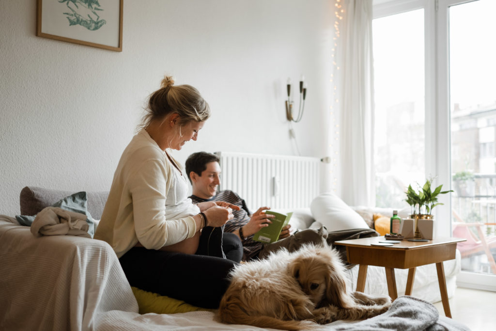 ein Mann und eine schwangere Frau sitzen auf einem Sofa in einem Wohnzimmer, die Frau ist schwanger, sie strickt, der Mann sitzt über Eck neben ihr, er blättert in einem Buch, neben der Frau sitzt ein Hund