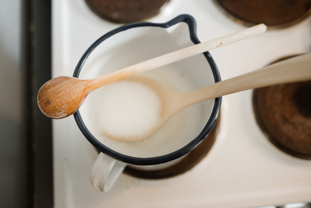in einem Emaille- Topf, der auf einem Herd steht, ist ein Holzkochlöffel und Milch, auf dem Topf iegt ein weiterer Holzkochlöffel