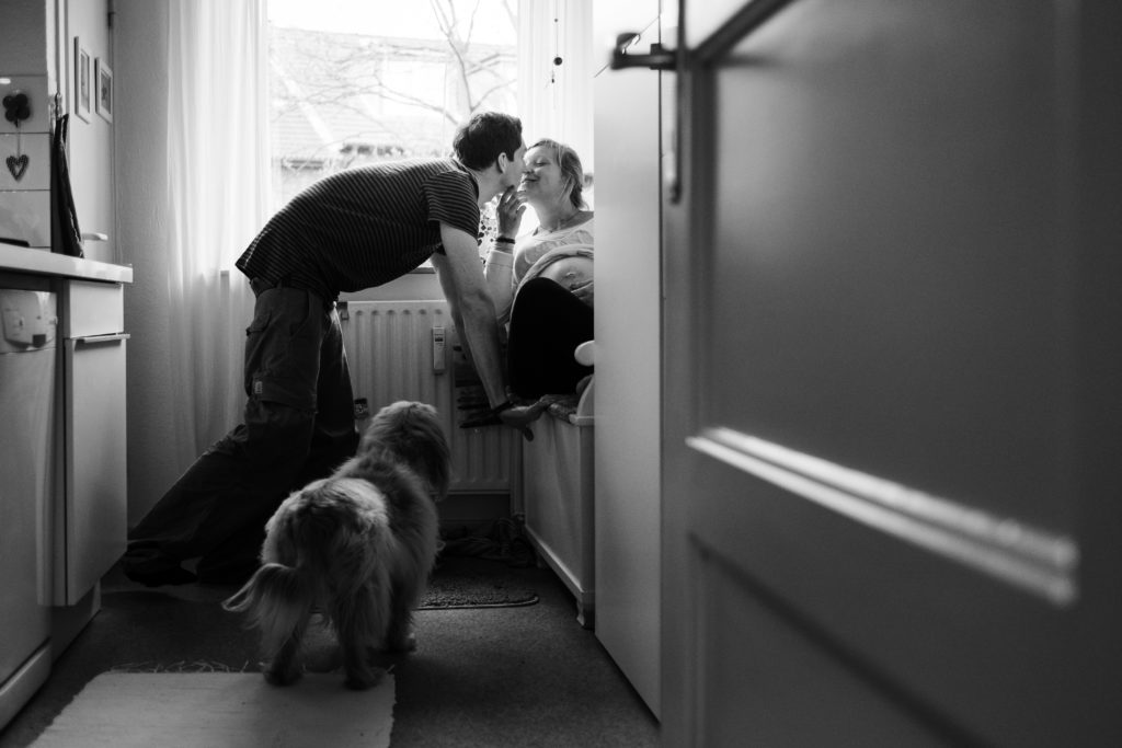 in einer Küche liegt eine schwangere Frau auf einer Holzbank, ein Mann steht neben der Bank und beugt sich zu ihr und gibt ihr einen Kuss, neben ihm steht ein Hund