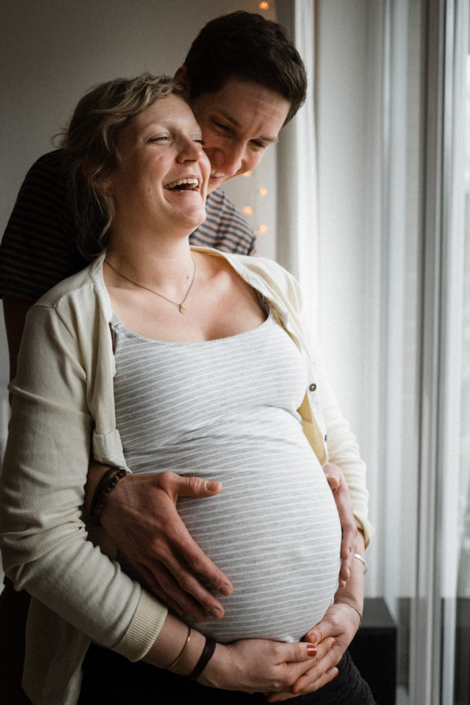 ein Mann und eine Frau stehen an einem Fenster in einer Wohnung, der Mann steht hinter der schwangeren Frau, er hält seine Hände an ihren Bauch, sie hält ihre Hände unter ihren Bauch, beide lachen