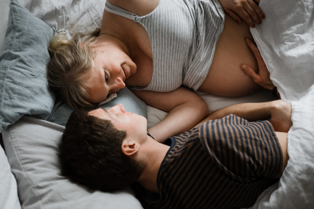 ein Mann und eine Frau liegen in einem Schlafzimmer in einem Bett, sie schauen sich gegenseitig an, der Mann legt seine Hand auf den Bauch der Frau, sie ist schwanger