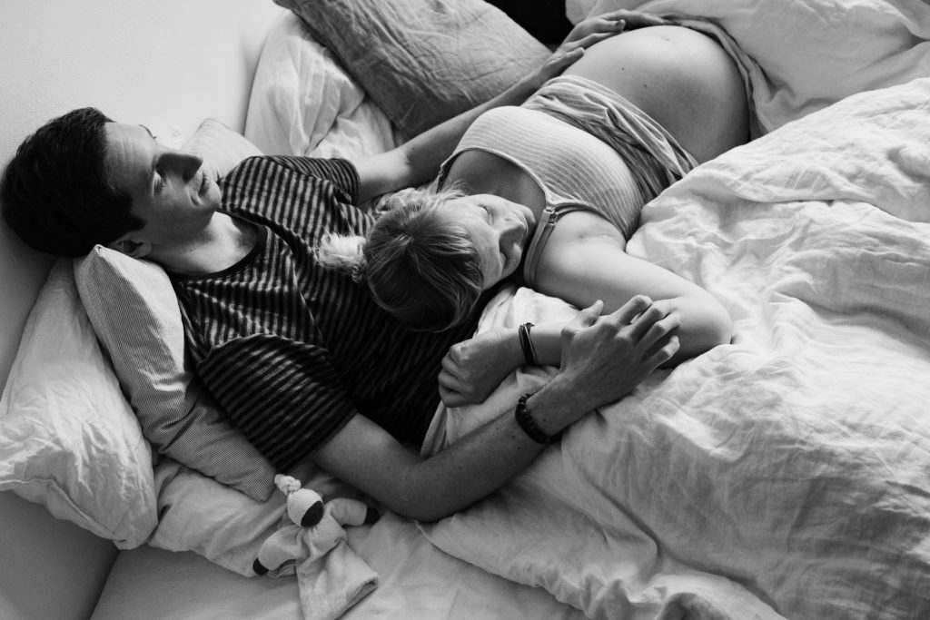 ein Mann und eine Frau iegen in einem Schlafzimmer in einem Bett, die Frau ist schwanger, sie legt ihren Kopf auf den Bauch des Mannes, die Hand des Mannes liegt auf den Bauch der Frau, beide schließen die Augen