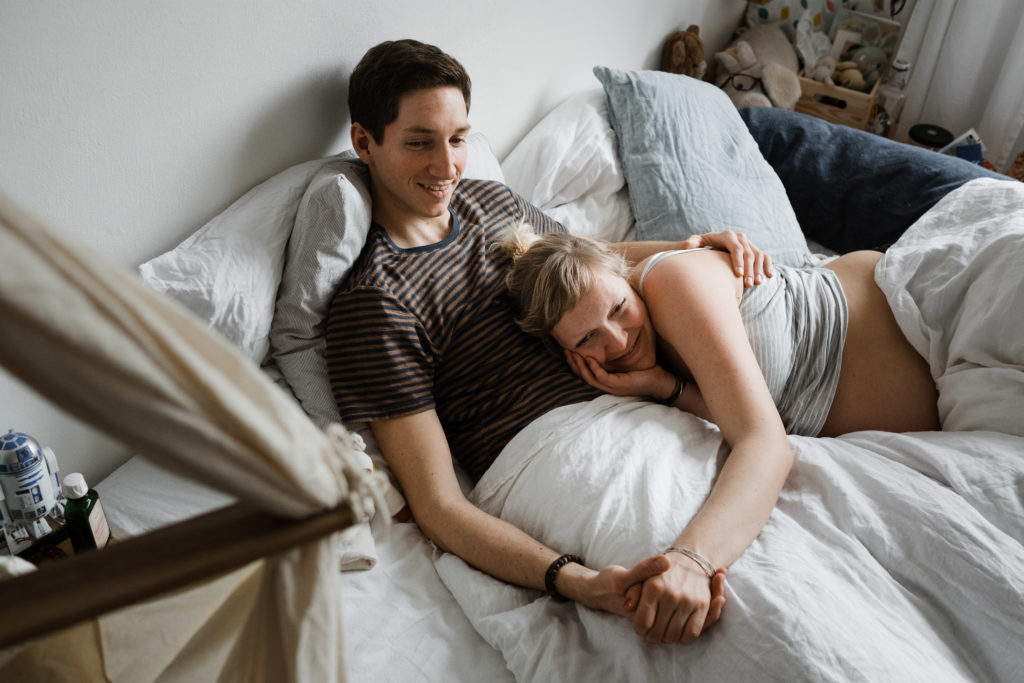 ein Mann und eine Frau iegen auf einem Bett in einem Schlafzimmer, die Frau ist schwanger, sie legt ihren Kopf auf den Bauch des Mannes, sie halten ihre Hand, sie schauen in die gleiche Richtung