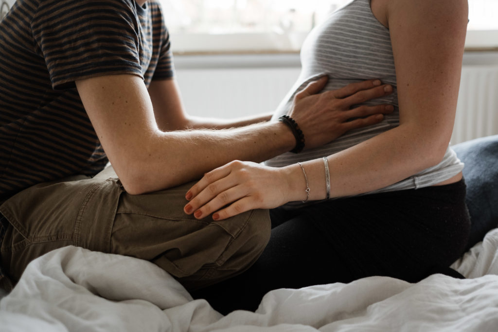 ein Mann und eine schwangere Frau sitzen sich in einem Bett in eienm Schlafzimmer gegenüber, der Mann hält seine Hände seitlich an den Bauch der Frau