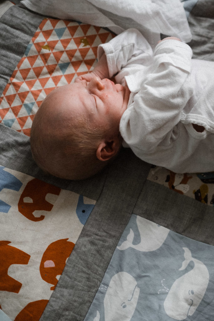 ein Baby liegt auf einer bunten Wickeldecke, es hat geschlossene Augen das Baby trägt einen weißen Body