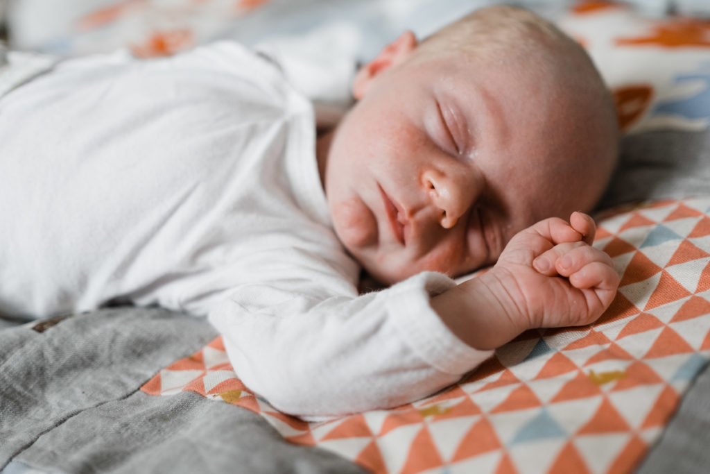 ein Baby liegt auf einer bunten Wickeldecke, es hat geschlossene Augen das Baby trägt einen weißen Body, die Arme des Baby sind nach oben gestreckt