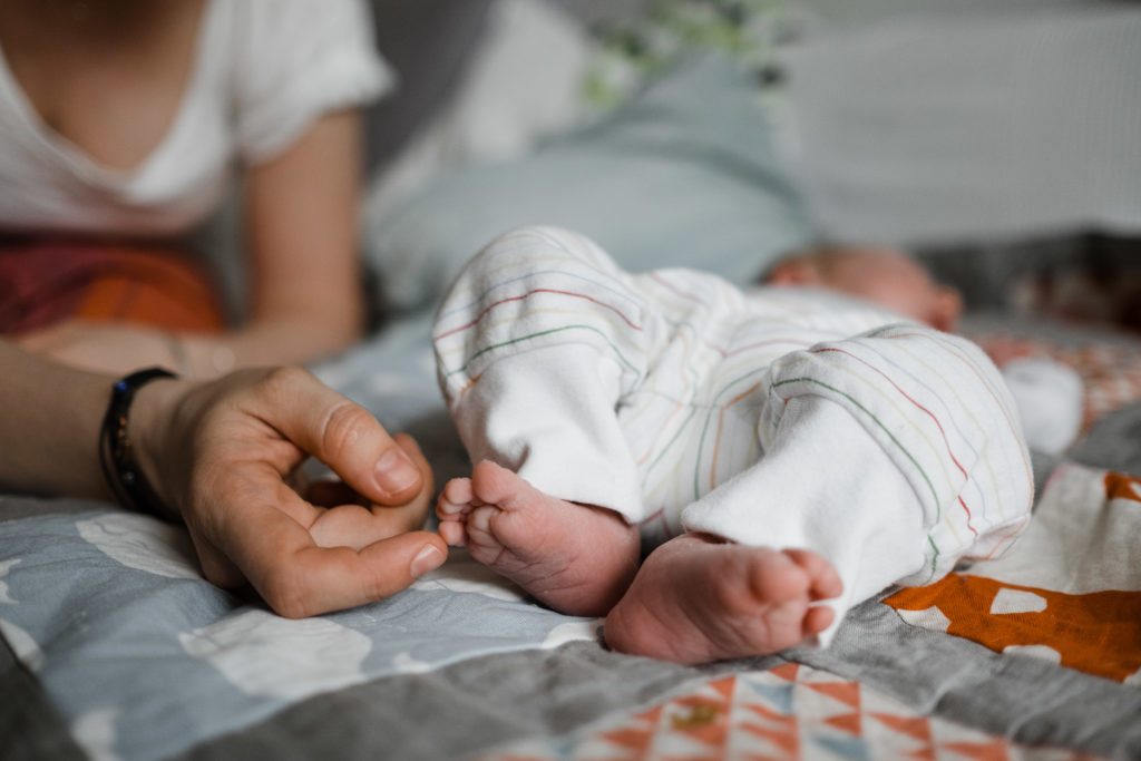 ein Baby liegt auf einer bunten Wickeldecke, das Baby trägt einen weißen Body, man sieht nur die Beine des Baby, eine Frau liegt neben dem Baby und hält ihre Hand an den Fuß des Baby
