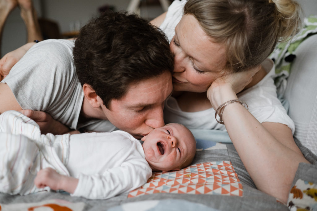 ein Mann und eine Frau liegen auf einem Sofa, vor ihnen liegt ein Baby auf einer Wickeldecke, das Baby hat einen weit geöffneten Mund, der mann küsst das Baby an der Wange, die Frau küsst den Man auf seine Schläfe