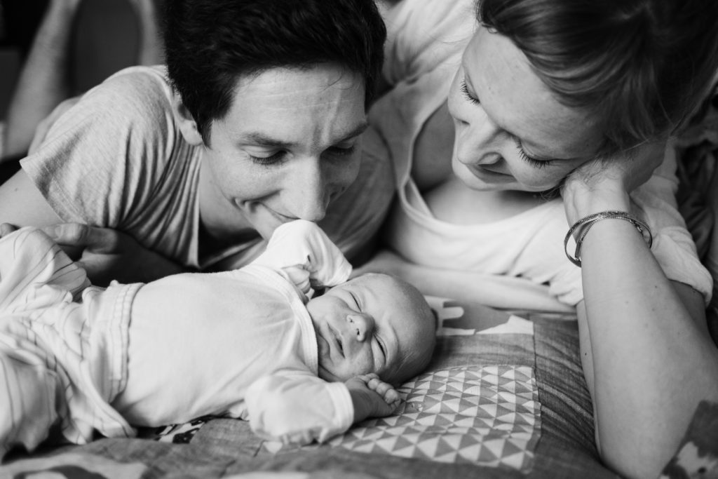 eine Mann und eine Frau liegen auf einem Sofa, vor ihnen liegt ein Baby auf einer Wickeldecke, das Baby streckt die Arme von sich, der Mann und die Frau schauen das Baby an