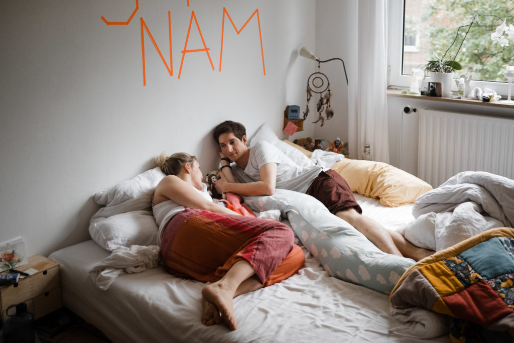 auf einem Bett im Schlafzimer liegen ein Mann und eine Frau, sie schauen sich an, zwischen ihnen liegt ein Baby