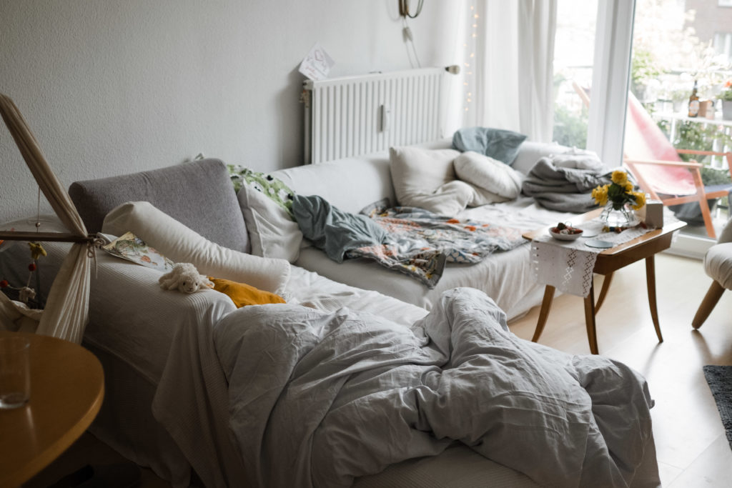 ein Sofa steht in einem Wohnzimmer, auf dem Sofa liegen verstreut Kissen und Decken, vor dem Sofa stht ein Beistelltisch mit einer Spitzendecke und einer Blumenvase