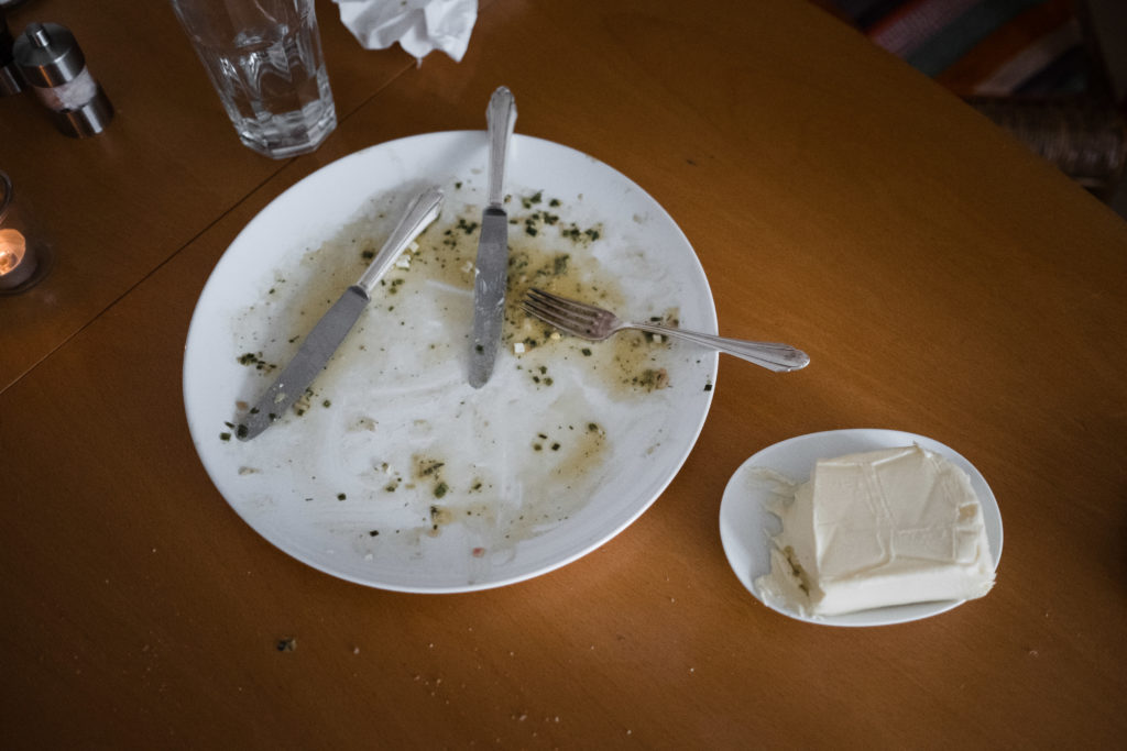 auf einem Holztisch steht ein Teller, auf dem gegessen wurde, das Besteckt liegt auf dem Teller, neben dem Teller steht ein Stück Frischkäse