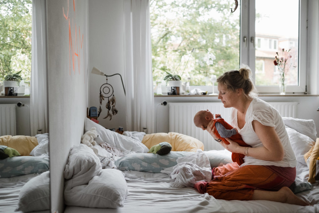 auf einem Bett im Schlafzimmer kniet eine Frau, sie hält ein Baby in den Armen, die Frau schaut das Baby an, neben ihr ist ein großes Fenster