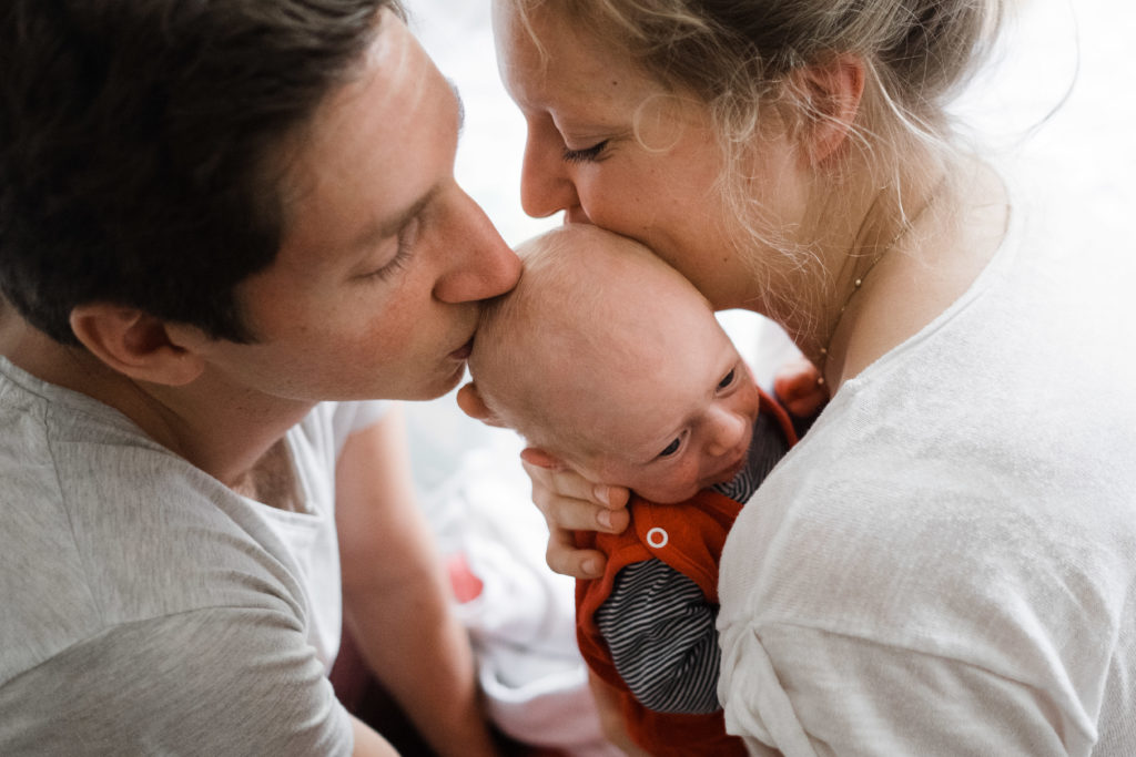 eine Frau hat ein Baby auf dem Arm, sie küsst das Baby auf dem Kopf, neben ihr steht ein Mann, der ebenfalls das Baby auf dem Kopf küsst