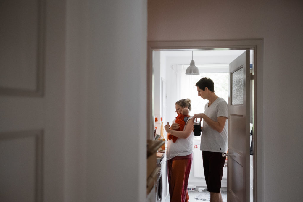 in einer Küche stehn ein Mann und eine Frau, die Frau hat ein Baby auf dem Arm, der Mann hat ene Dose in der Hand, der Mann schaut auf die Frau