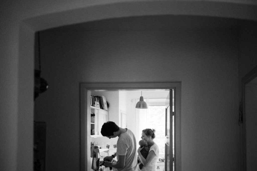 ein Mann und eine Frau stehen in einer Küche, die Frau hat ein Baby auf dem Arm, der Mann steht an der Küchenzeile