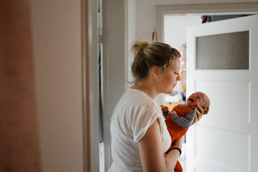 eine Frau steht in einem Flur, sie hat ein weinendes Baby auf den Arm, die hält den Kopf des Baby mit hrer Hand