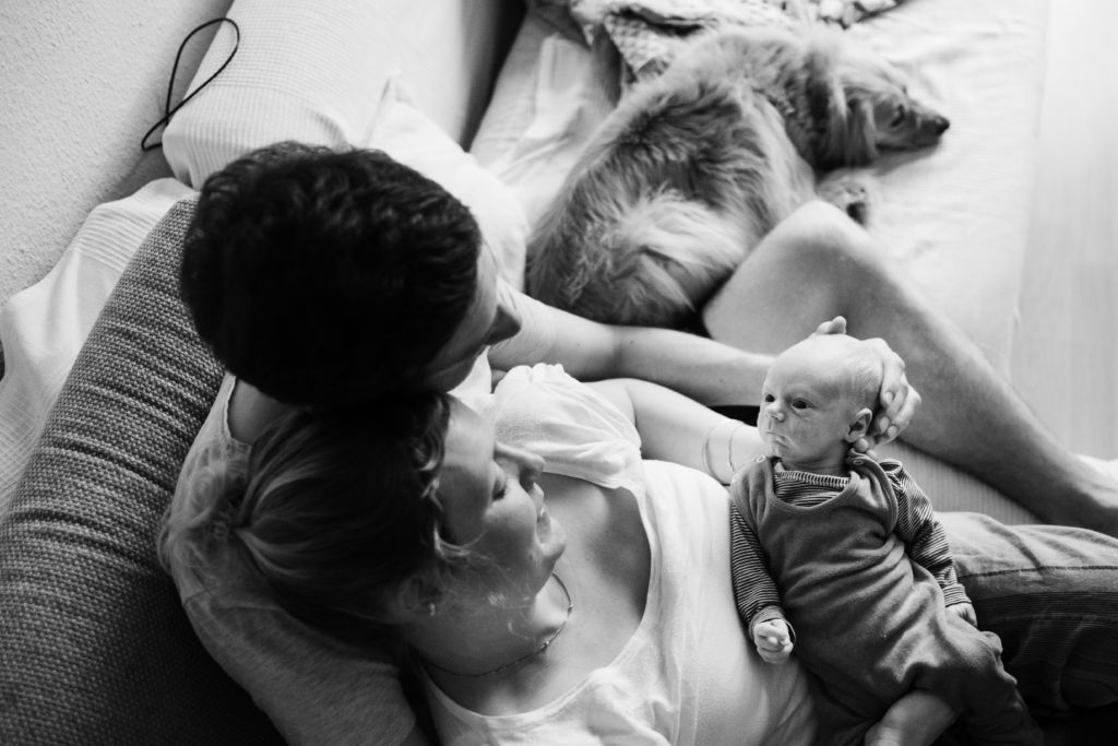 ein Mann und eine Frau sitzen auf einem Sofa im Wohnzimmer, die Frau sitzt in den Armen des Mannes, sie hält ein Baby in ihren Händen, beide schauen auf das Baby, neben ihnen liegt ein Hund mit auf dem Sofa