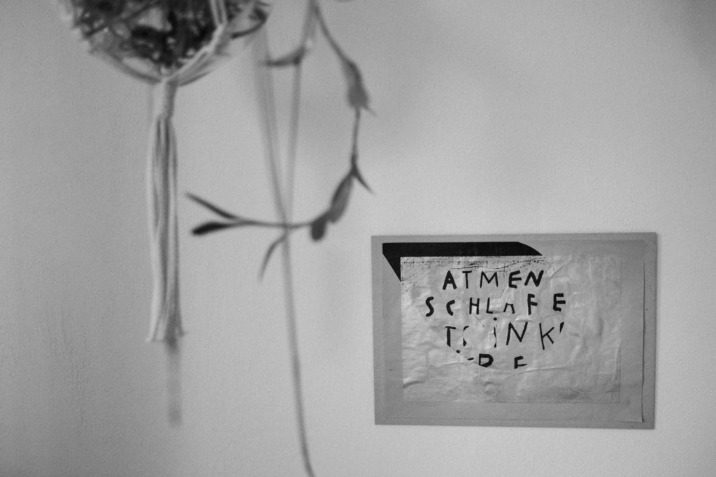 vor einer weißen Wan dhängt ein Bild mit einer schwarz/ weißen Schrift, im Vordergrun hängt eine Makramee- Blumenampel