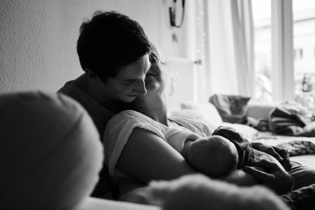 ein Mann und eine Frau sitzen auf einem Sofa im Wohnzimmer, der Mann sitzt hinter der Frau, die Frau hat ein Baby auf dem Arm, sie neigt ihren Kopf nach hinten an den Kopf des Mannes, der Mann schaut auf das Baby