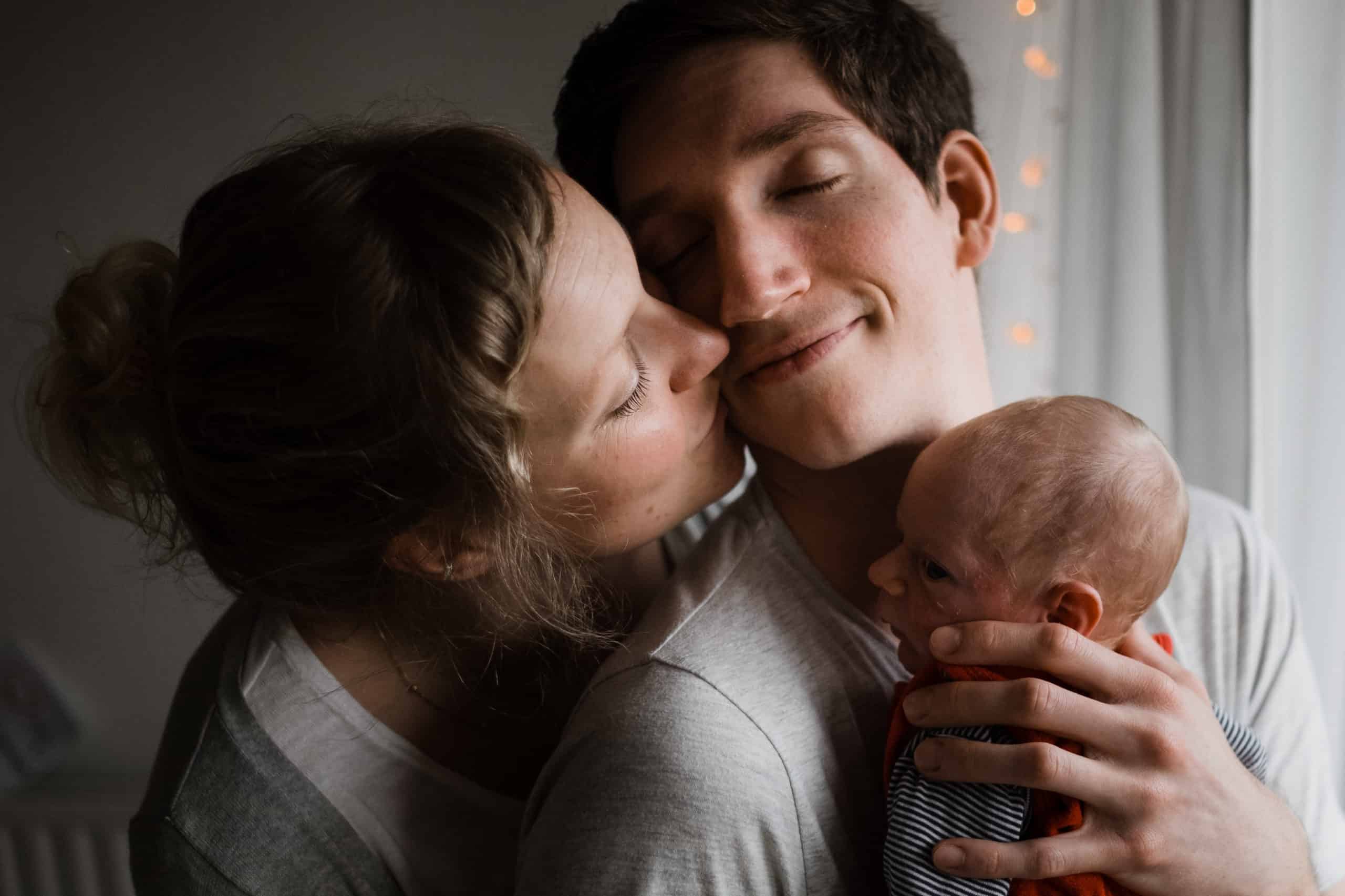 vor einem Fenster in einer Wohnung steht ein Mann, er hat ein Baby auf dem Arm, er hält seine Hand an den Kopf des Baby, er neigt seinen Kopf zu der Frau, die neben ihm steht, die Frau küsst ihn neben seinen Mund, beide haben geschlossene Augen