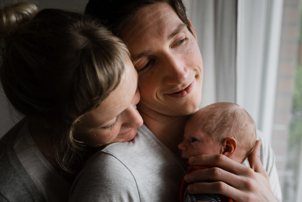 vor einem Fenster in einer Wohnung steht ein Mann, er hat ein Baby auf dem Arm, er hält seine Hand an den Kopf des Baby, er schaut aus dem Fenster, neben ihm steht eine Frau, die sich auf seine Schulter lehnt, sie schaut auf das Baby