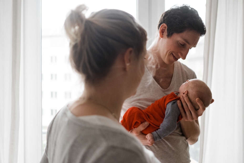 ein Mann steht mit dem Rücken zu einem Fenster in einer Wohnung, er hat ein Baby auf dem Arm, er hält seine Hand unter den Kopf des Baby und schaut zu ihm, neben ihm steht eine Frau, die auf das baby schaut