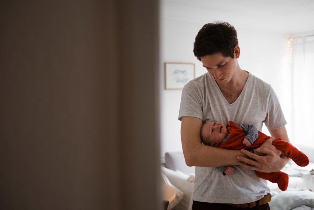 ein Mann steht in der Tür zum Wohnzimmer, er hat ein Baby auf dem Arm, das Baby weint, der Mann schaut hinunter auf das Baby