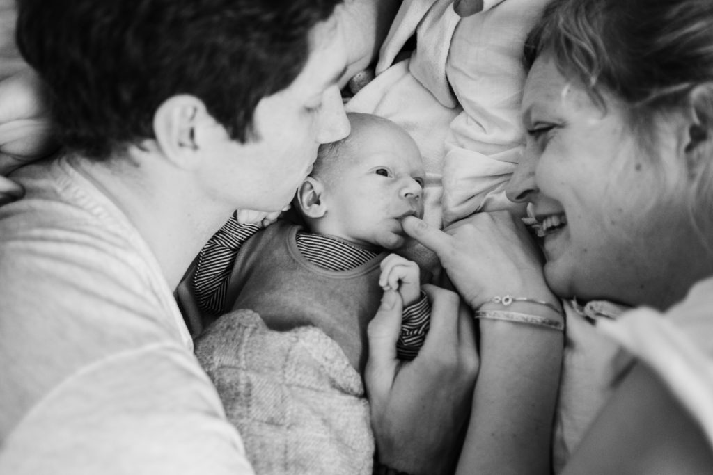 auf einem Bett im Schlafzimmer liegen ein Mann und eine Frau, zwischen ihnen liegt ein Baby, der Mann und die Frau schauen das Baby an , die Frau hält ihren kleinen Finger an den Mund des Baby