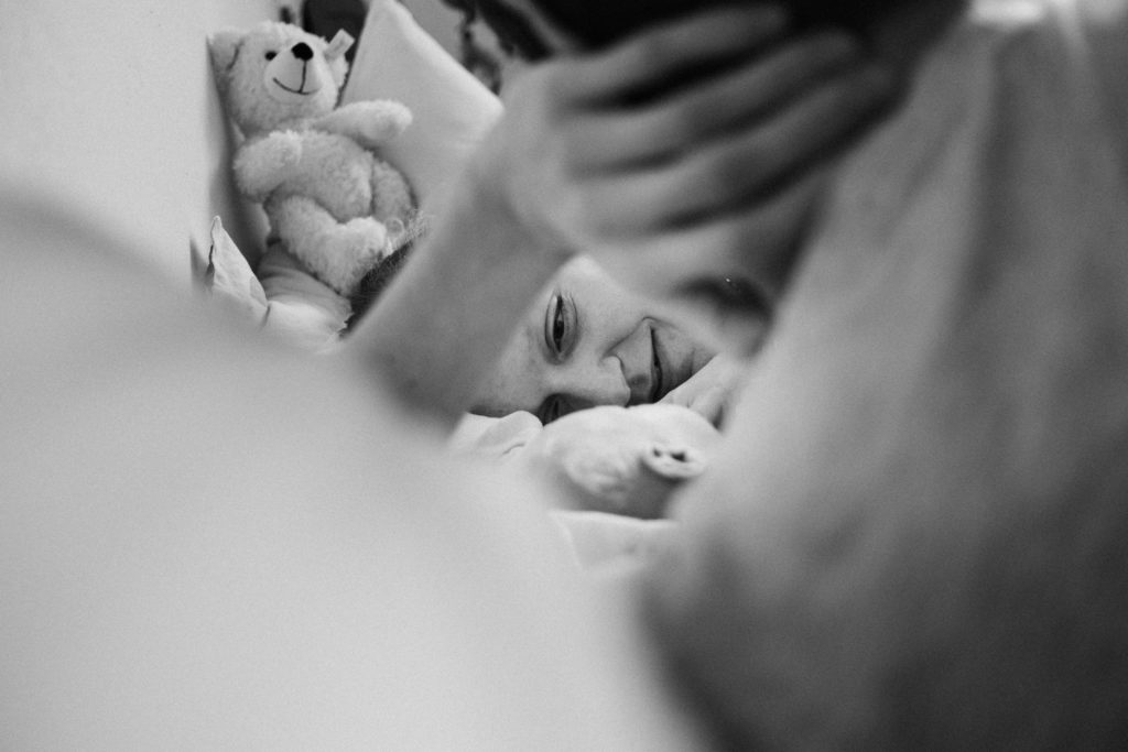 ein Mann und eine Frau liegen auf einem Bett im Schlafzimmer, zwischen ihnen liegt ein Baby, der Mann und die Frau schauen das Baby an, zu sehen ist nur ein Ausschnitt der Gesichter