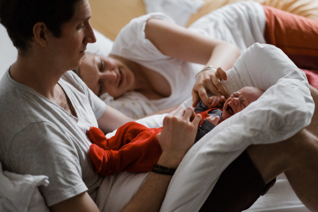 auf einem Bett im Schlafzimmer liegt eine Frau, neben ihrsitzt ein Mann mit aufrechten Beinen, auf seinen Beinen liegt auf einem Kissen ein Baby, es weint, die Frau hält ihre Hand an die Hand des Baby