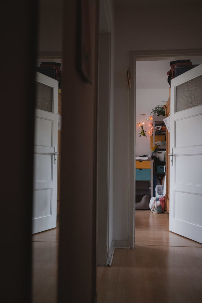 in einem Flur stehen einige weiße Zimmertüren offen, durch eine Tür sieht man eine bunte Lichterkette und ein Holzregal