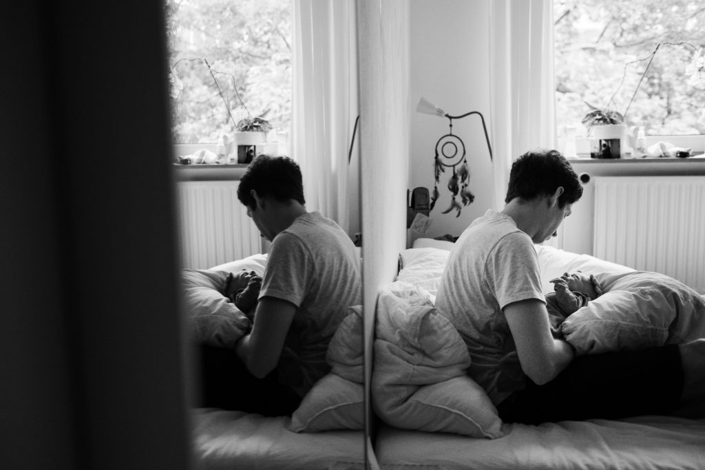 auf einem Bett im Schlafzimmer sitzt ein Mann, der ein Baby in dem Armen hält, er schaut zu dem Baby und hält seine Hand unter den Kopf des Baby, man sieht eine Spiegelung in dem Spiege