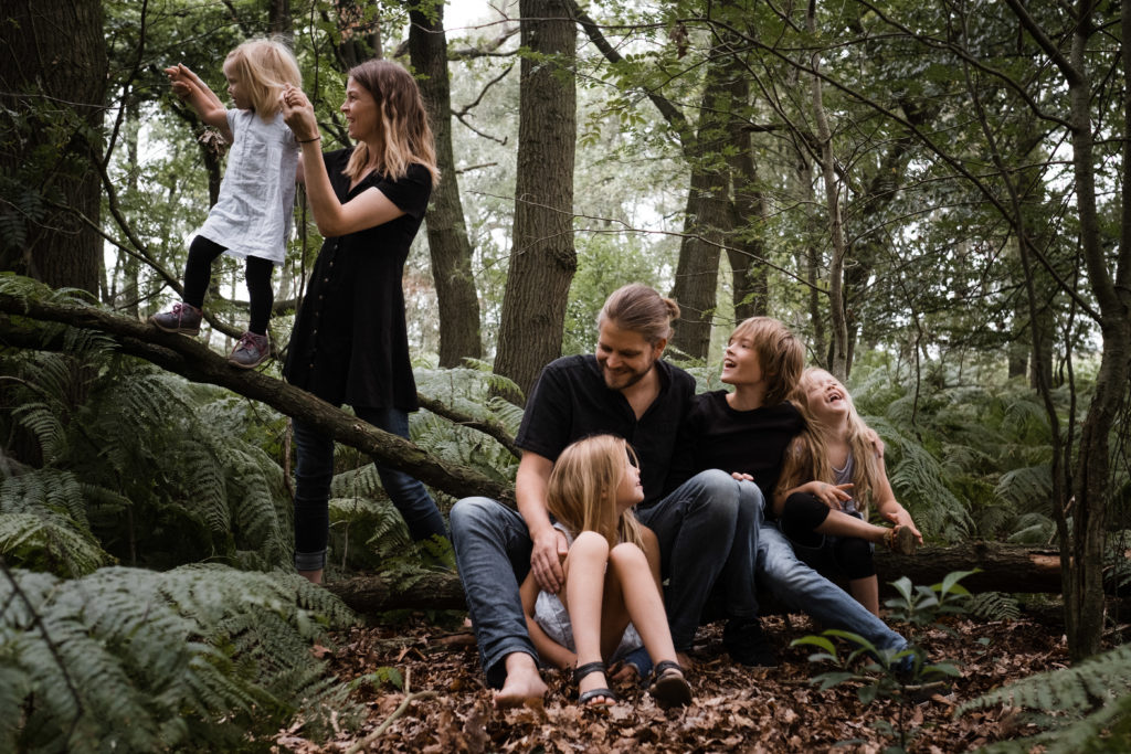 im Wald sitzt ein Vater mit seinen 2 Töchtern und seinem Sohn auf einem Baumstamm, die Mutter hält ihre Tochter, sie balanciert auf dem Baumstamm