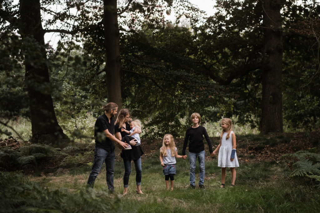 die Familie steht in einem Wald nebeneinander, die Mutter trägt die kleinste Tochter, ihre beiden anderen Töchter und der Sohn stehen daneben, alle drei halten sich an den Händen, der Vater steht neben seiner Frau