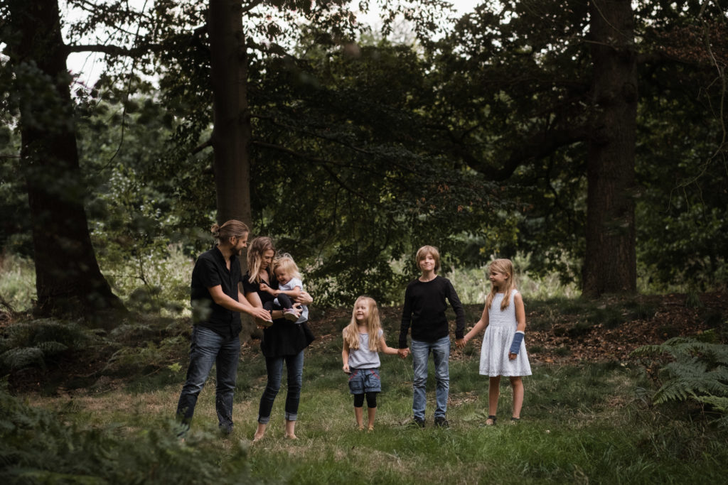 die Familie steht in einem Wald nebeneinander, die Mutter trägt die kleinste Tochter, ihre beiden anderen Töchter und der Sohn stehen daneben, alle drei halten sich an den Händen, der Vater steht neben seiner Frau