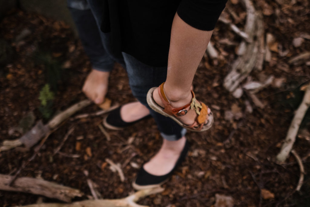 eine Mutter hat ihre Tochter auf dem Arm, die Beine der Tochter hängen, man sieht nur die Beine der beiden, sie stehen auf Waldboden