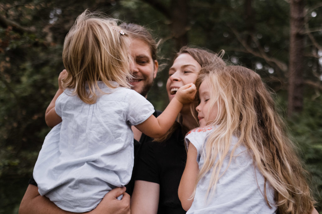 ein Vater hat seine Tochter auf dem Arm, daneben steht die Mutter, die auch ihre Tochter auf dem Arm hat, sie schauen sich gegenseitig an, sie stehen im Wald
