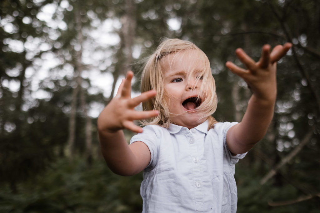ein Mädchen streckt die Arme nach vorne, ihre Finger spreizt sie, sie lacht, der Wind weht ihre Harae ins Gesicht, sie steht im Wald