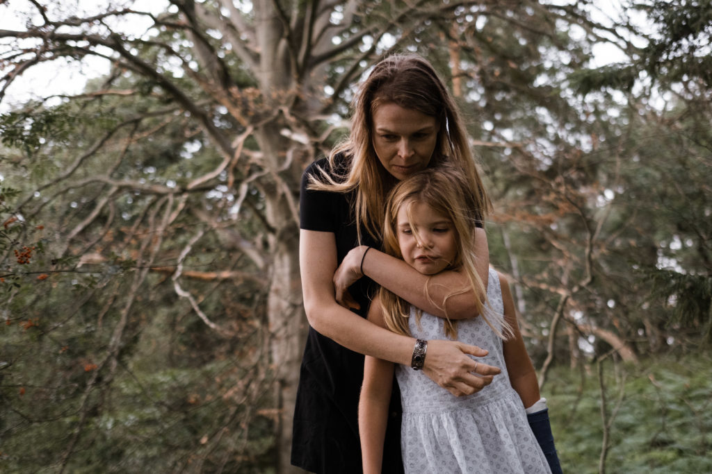 eine Mutter hält ihre Tochter an ihren Körper, sie stützt ihren Kopf auf den Kopf ihrer Tochter, sie stehen im Wald vor einem großen Baum