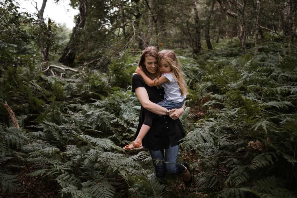 eine Mutter trägt ihre Tochter auf dem Arm durch einen Wald, der Boden ist mit großem Fahn bewachsen, die Tochter hält sich am Hals der Mutter fest